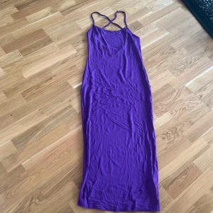 Lila långklänning med smala axelband - Snygg lila långklänning med smala korsade axelband och öppen rygg. Perfekt för en elegant kvällslook. Klänningen har en figurnära passform som framhäver silhuetten.