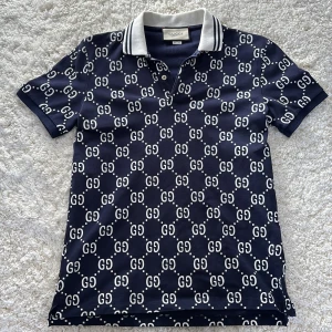 Mörkblå pikétröja från Gucci - Säljer en stilren mörkblå pikétröja från Gucci med det ikoniska GG-mönstret i vitt. Tröjan har en klassisk krage med vita och svarta ränder och korta ärmar. Perfekt för en trendig look. Är inte alls använd mycket, ett fåtal gånger.