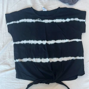 Svart och vit randig t-shirt från Lindex - Säljer en cool svart t-shirt från Lindex med vita ränder i tie-dye stil. T-shirten har korta ärmar och en knytning framtill som ger en avslappnad look. Perfekt för en trendig vardagsstil!