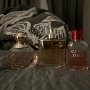 Parfymflaskor - Tre olika parfymer till salu. Första flaskan är rund med en guldig kork och har en ljusrosa vätska och doftar rosor. Andra flaskan är fyrkantig med en transparent kork och innehåller en gyllene vätska och doftar som La Vie est Belle.  Tredje flaskan är från La Rive, heter 'Sweet Rose', och har en röd kork med en rosa vätska.