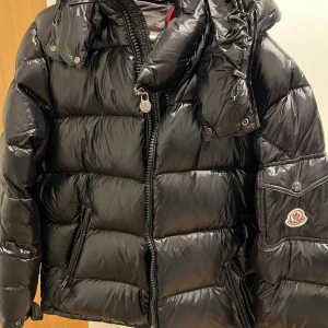 Svart pufferjacka från Moncler - Säljer en snygg svart pufferjacka från Moncler. Jackan har en glansig finish och är utrustad med dragkedja och huva. Perfekt för kyliga dagar när du vill hålla stilen. Moncler-logga på ärmen ger en exklusiv touch.