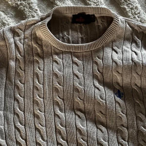 Grå kabelstickad tröja från morris  - Säljer en stilren grå kabelstickad tröja från Morris med rund halsringning. Tröjan har ett diskret broderat märke på bröstet i blått. Perfekt för kyligare dagar och ger en klassisk look.