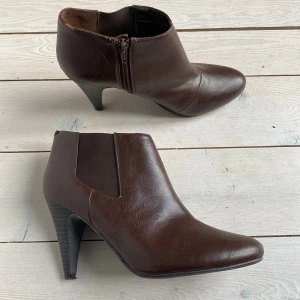 Bruna boots med klack - Låga bruna boots/stövletter i konstskinn med spetsig tå och dragkedja på sidan och resår på andra. Ena skon har en repa på klacken och små skador i övre kanten. Se bild. Storlek 38 men lite smala i tån. Från H&M. Använda ett fåtal gånger. 