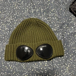 Gp company mössa - Snygg olivgrön mössa med ribbad design och coola glasögonliknande detaljer framtill. Perfekt för att ge din outfit en unik touch. Passar bra för kyligare dagar.