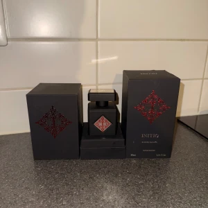 Initio Blessed Baraka - Elegant parfym från Initio i en stilren svart flaska med röda detaljer. Flaskan rymmer 90 ml och kommer i en matchande svart kartong med dekorativa röda mönster. Perfekt för den som söker en lyxig doftupplevelse.