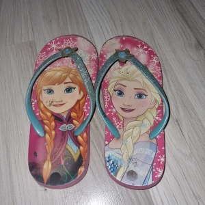 Rosa sandaler med Frost-motiv - Säljer ett par rosa sandaler med motiv av karaktärer från Frost. Sandalerna har glittriga remmar och en liten blomdetalj framtill. Perfekta för sommarens äventyr!