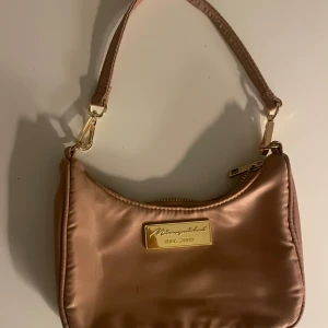 Rosé axelväska med gulddetaljer - Snygg rosé liten axelväska med gulddetaljer och dragkedja. Väskan har en elegant design med en justerbar axelrem och en logotypplatta framtill. Perfekt för att bära dina nödvändigheter med stil.