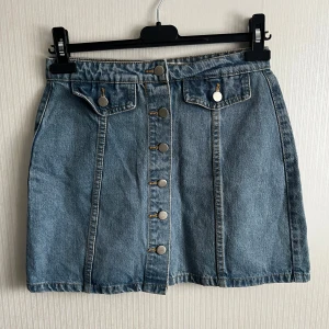 Jeanskjol från Denim Co. - Snygg jeanskjol från Denim Co. i klassisk blå denim. Kjolen har knappar framtill och två dekorativa fickor med knappar. Perfekt för en avslappnad stil.
