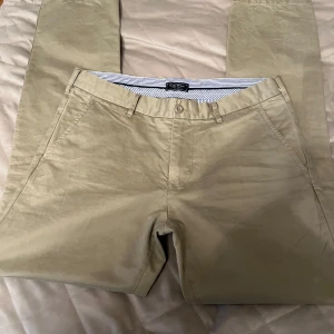 Beige chinos från East West - Snygga beige chinos från East West i 100% bomull. De har en klassisk design med knappar och bälteshällor. Perfekta för en stilren look.