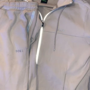 Ljusgrå tracksuit från Boss - Säljer en stilren beige tracksuit från Boss med dragkedja och matchande byxor. Tröjan har en diskret logga på bröstet och byxorna har en elastisk midja med logga. Perfekt för en avslappnad stil.