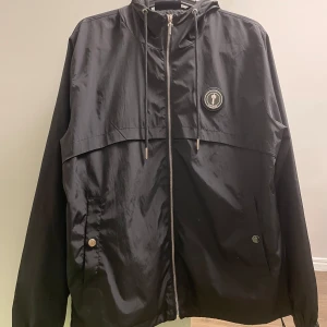 Svart windbreaker från Trapstar - Snygg svart vindjacka från Trapstar med dragkedja och huva. Jackan är i bra kondition