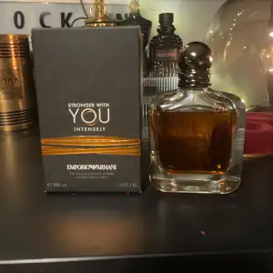 Säljer en flaska Stronger With You Intensely Eau de Parfum från Emporio Armani. Flaskan är elegant med en mörk bärnstensfärgad vätska och en stilren design. Förpackningen är svart med gyllene detaljer och text. Perfekt för den som vill ha en intensiv och sofistikerad doft. (Boxen ingår ej)
