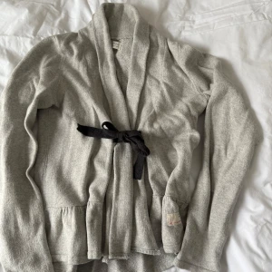 Grå kofta från Odd Molly - Säljer en mysig grå kofta från Odd Molly i bomull. Koftan har långa ärmar och ett mörkgrått knytband i midjan. Den har en broderad detalj på ryggen och en volangkant nedtill. Perfekt för kyliga dagar!