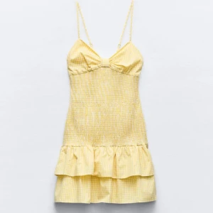 Gul rutig klänning med volang - Söt gul klänning med rutigt mönster och volangdetalj. Axelbandslös design med smala axelband och kort längd. Perfekt för sommarens varma dagar.