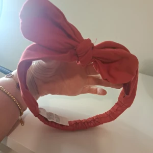 Rött diadem med rosett - Snyggt rött diadem med en stor rosett framtill. Perfekt för att ge en färgklick till din outfit. Diademet har en elastisk del för bättre passform.
