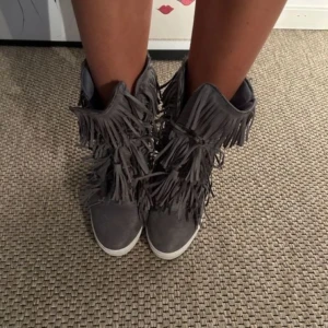 Fransskor - Snygga grå mockaboots med fransar och snörning. Dessa boots har en diskret kilklack och är perfekta för att ge din outfit en bohemisk touch. OBS första bilden är lånad