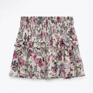 Blommig kortkjol med volang - Söt blommig kortkjol med volangdetaljer i rosa och svart på en vit bas. Perfekt för sommarens varma dagar. Kjolen har en elastisk midja för en bekväm passform. Storlek m men passar xs-m