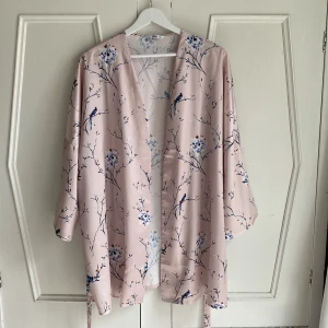 Rosa kimono med fågelmönster - Säljer en elegant blekrosa kimono med ett vackert fågel- och blommönster i blått och vitt. Kimonon har trekvartsärmar och ett knytband i midjan för en justerbar passform. Perfekt för en stilfull och avslappnad look. Aldrig använd. 
