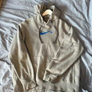 Grå hoodie från Nike - Säljer en grå hoodie från Nike med en blå logga på bröstet. Luvan är justerbar. 