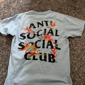 Ljusblå t-shirt från Anti Social Social Club - Säljer en ljusblå t-shirt från Anti Social Social Club med stort tryck på baksidan och mindre logga på framsidan. T-shirten har korta ärmar och är perfekt för en avslappnad stil.