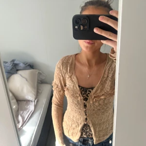 Beige spetskofta med knappar - Säljer en elegant beige spetskofta med knappar framtill. Koftan har ett vackert spetsmönster och långa ärmar med volangdetaljer vid ärmsluten. Perfekt för att lägga till en romantisk touch till din outfit.