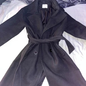 Svart kappa med bälte - Snygg svart kappa från Bershka med bälte i midjan och knappar framtill. Perfekt för kyligare dagar med sin klassiska design och långa ärmar. Ett måste i garderoben för en stilren look.