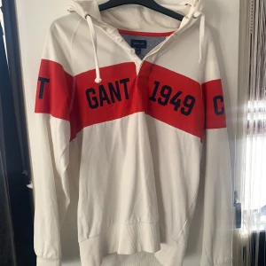 Vit och röd hoodie från GANT - Säljer en snygg vit hoodie från GANT med röda detaljer och texten '1949' och 'NEW HAVEN'. Hoodien har en justerbar huva med snören och knappar framtill. Perfekt för en avslappnad stil.