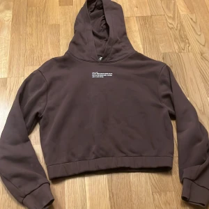 Brun croppad hoodie från H&M - Säljer en brun croppad hoodie från H&M med text på bröstet. Hoodien har långa ärmar och en bekväm passform. Perfekt för en avslappnad stil.