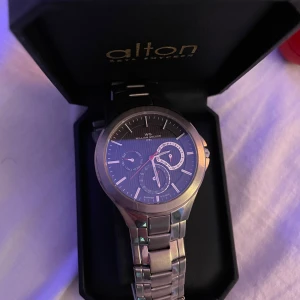 Silverfärgad klocka från Alton - Snygg och stilren klocka från Alton med silverfärgad metallarmband och mörk urtavla. Klockan har flera små urtavlor och tydliga markeringar. Perfekt för den som gillar en klassisk look.