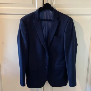 Mörkblå kavaj från Dressmann - Snygg mörkblå kavaj från Dressmann i storlek 46. Kavajen har en klassisk design med två knappar framtill och en stilren passform. Perfekt för både formella och semi-formella tillfällen. Fodret är i en ljusare nyans med röda detaljer. Endast använd 1 gång. 