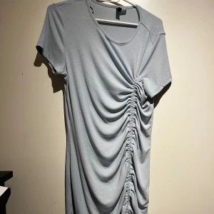 Ljusblå klänning med rynkdetalj - Snygg ljusblå klänning med korta ärmar och en cool rynkdetalj längs sidan som ger en unik look. Perfekt för en avslappnad stil med en twist. Klänningen har en rund hals och är gjord i ett mjukt material för extra komfort.