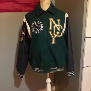 Grön och svart bomberjacka med Bronx Bears-märke - Snygg grön och svart bomberjacka med broderade detaljer och stort 'NYC' märke på framsidan. Jackan har långa ärmar och ribbade muddar. Perfekt för en sportig look!