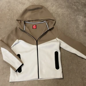 Beige och vit tröja från Nike - Säljer en snygg beige och vit tröja från Nike med huva och dragkedja. Tröjan har långa ärmar och svarta detaljer vid fickorna. Perfekt för en sportig look! 2 ställen där det finns lite skit men inte något märkvärdigt. Är cirka 1,65 och den sitter perfekt.