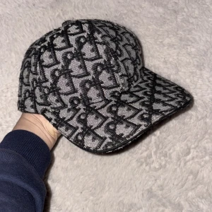 Grå och svart mönstrad keps från dior - Snygg keps med ett unikt mönster i grått och svart. Perfekt för att ge din outfit en cool touch. Kepsen har en klassisk skärm och är tillverkad i ett slitstarkt material.
