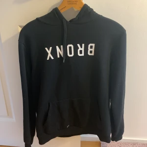 Svart hoodie med text - Säljer en svart hoodie med texten 'BRONX' i vitt på framsidan. Tröjan har en klassisk design med huva och känguruficka. Perfekt för en avslappnad stil.