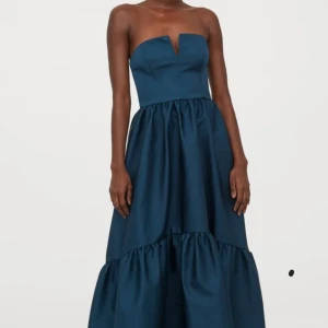 Mörkblå axelbandslös klänning - Hm Sudio collection. Elegant mörkblå axelbandslös klänning med volangdetaljer. Klänningen har en lång och flödande design, perfekt för speciella tillfällen. Den är tillverkad i ett glansigt tungt material som ger en lyxig känsla.