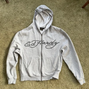 Grå hoodie från Ed Hardy - Säljer en grå hoodie från Ed Hardy i samarbete med Urban Outfitters. Den har en dragkedja framtill och ett stort tigertryck på huvan. Perfekt för en avslappnad stil.