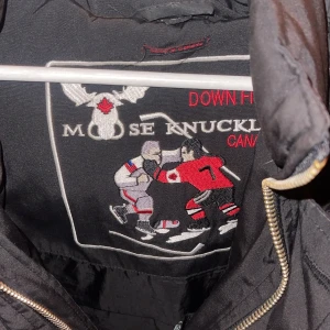 Svart dunjacka från Moose Knuckles - Säljer en svart dunjacka från Moose Knuckles med pälskrage och broderad logga på insidan. Jackan har dragkedja och knappar framtill samt en ficka med dragkedja på ärmen. Perfekt för kalla dagar!