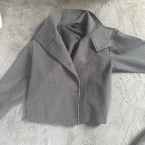 Grå kappa med bred krage - Snygg grå kappa med bred krage och knappar framtill. Perfekt för höst och vinter. Den har en stilren design och är tillverkad i ett mjukt material som ger en elegant look.