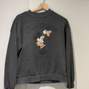 Grå sweatshirt från Disney - Säljer en grå sweatshirt från Disney med ett tryck av Musse Pigg och texten 'True Original'. Tröjan har långa ärmar och en rund halsringning. Perfekt för alla Disney-fans! 🐭
