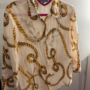 Antica Sartoria blus/skjorta - Säljer en unik vit blus från Antica Sartoria med ett iögonfallande repmönster i brunt och detaljer av paljetter. Blusen har långa ärmar och knappar framtill. Perfekt för dig som vill sticka ut med en stilren men ändå lekfull look.