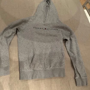 Grå hoodie från Tommy Hilfiger - Säljer en klassisk grå hoodie från Tommy Hilfiger med logotyp på bröstet. Tröjan har en bekväm passform med långa ärmar och en praktisk magficka. Perfekt för en avslappnad stil.