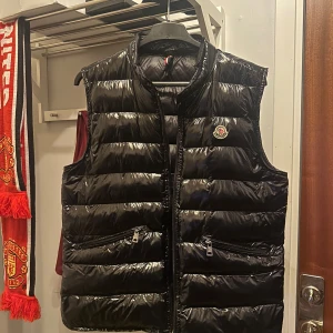 Svart dunväst från Moncler - Säljer en snygg svart dunväst från Moncler med glansig yta. Västen har två praktiska fickor med dragkedjor och Moncler-logga på bröstet. Dragkedja trasig så går inte att stänga, går säkert lätt att lösa om man vill det!