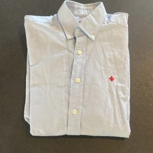 Ljusblå skjorta från Morris - Snygg ljusblå skjorta från Morris med ett  rött broderat märke på bröstet. Andledning är att den är för liten. Knappt andvänd väldigt stilren.                         Nypris är 1199 men mitt pris är bara 250!!   Hör av dig vid fler frågor eller om mer bilder begärs! Storlek 37 passar S 