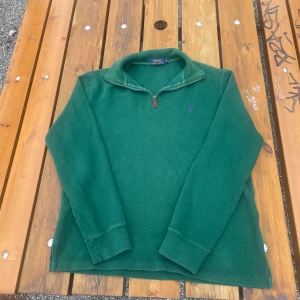 Grön tröja med dragkedja från Polo Ralph Lauren - Säljer en snygg grön tröja från Polo Ralph Lauren med dragkedja och lila broderad logga på bröstet. Perfekt för en stilren och avslappnad look. Tröjan har långa ärmar och en bekväm passform.