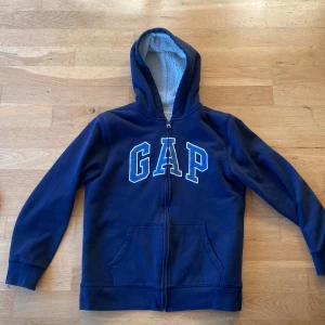 Mörkblå hoodie från GAP - Säljer en mörkblå hoodie från GAP med dragkedja och stor logga på framsidan. Tröjan har en mysig fodrad insida och långa ärmar. Perfekt för kyliga dagar!