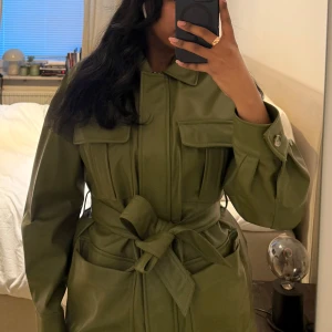 Grön skinnjacka med knytbälte - Snygg grön skinnjacka med knytbälte i midjan och knappar framtill. Jackan har långa ärmar och flera praktiska fickor. Perfekt för att ge en cool touch till din outfit.