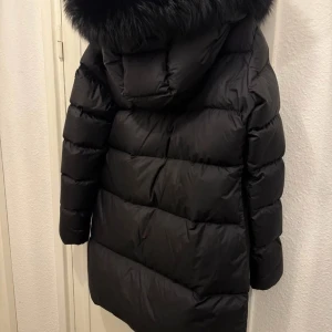 Svart dunjacka med pälshuva - Snygg svart dunjacka med en stor, fluffig pälshuva. Jackan har en lång passform och dragkedja framtill. Perfekt för kalla vinterdagar.