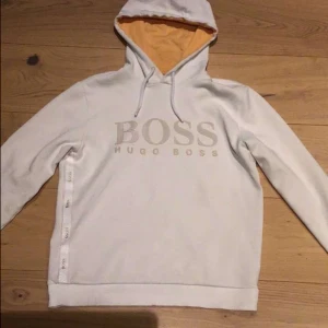 Vit hoodie från Hugo Boss - Säljer en stilren vit hoodie från Hugo Boss med logotypen tryckt på framsidan. Hoodien har en kontrasterande orange insida i huvan och ribbade muddar. Perfekt för en avslappnad stil.