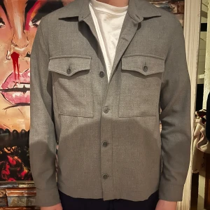 Grå overshirt  - Snygg grå overshirt med knappar framtill och två bröstfickor med lock. Perfekt för lager-på-lager-stil och passar till många olika outfits. Modellen är 190cm och bär Medium 